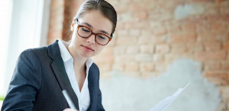 Young pretty accountant in eyeglasses and suit writing down her working ideas in notebook